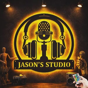 Puede incluir: Un letrero de metal negro con retroiluminación amarilla, que presenta un diseño de micrófono y auriculares. El letrero dice "JASON'S STUDIO" en letras blancas. Incluye una cámara, un altavoz y un mando a distancia.