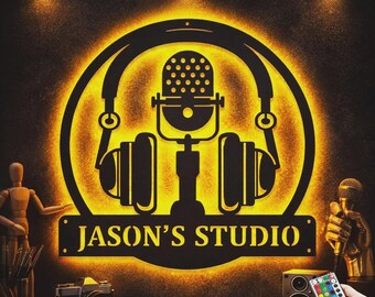 Może przedstawiać: Czarny metalowy znak z żółtym podświetleniem, przedstawiający mikrofon i słuchawki. Znak zawiera napis "JASON'S STUDIO" białymi literami. Dodatkowo aparat fotograficzny, głośnik i pilot.