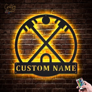 Könnte beinhalten: Ein schwarzes Metallschild mit einem runden Design, das zwei gekreuzte Hockeyschläger und einen Puck zeigt. Das Schild ist mit warmen gelben Lichtern beleuchtet und hat ein anpassbares Namensschild mit der Aufschrift "CUSTOM NAME".