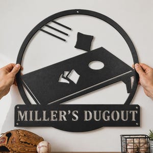 Könnte beinhalten: Schwarzes Metallschild mit einem Cornhole-Spiel-Design und dem Text "MILLER'S DUGOUT". Das runde Design zeigt ein Cornhole-Brett, Beanbags und ein Baseball-Thema. Ein Baseballhandschuh und ein Ball sind darunter zu sehen.