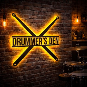 Puede incluir: Un letrero de metal negro con el texto "DRUMMER'S DEN" y baquetas cruzadas, iluminado con un cálido resplandor amarillo. El letrero está montado en una pared de ladrillo, creando un ambiente rústico.