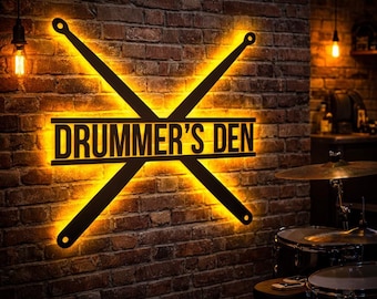 May include: A black metal sign, featuring the words "DRUMMER'S DEN" and crossed drumsticks, illuminated with a warm yellow light. The sign is mounted on a brick wall, creating a rustic atmosphere.