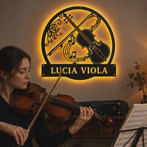Puede incluir: Un letrero de pared circular de metal negro con un dise&ntilde;o recortado de un viol&iacute;n y notas musicales, iluminado por una luz de fondo amarilla c&aacute;lida. El letrero incluye una base rectangular horizontal con el texto LUCIA VIOLA en letras may&uacute;sculas.