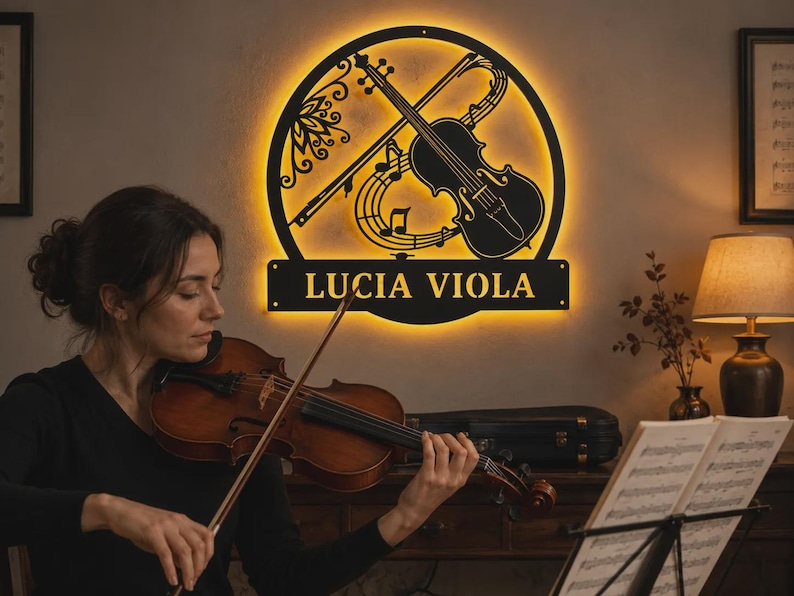 Puede incluir: Un letrero de pared circular de metal negro con un dise&ntilde;o recortado de un viol&iacute;n y notas musicales, iluminado por una luz de fondo amarilla c&aacute;lida. El letrero incluye una base rectangular horizontal con el texto LUCIA VIOLA en letras may&uacute;sculas.