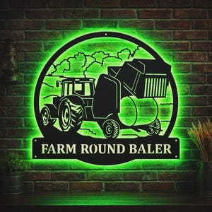 Può includere: Un'insegna da parete circolare in metallo nero con la sagoma di un trattore che traina una rotopressa, illuminata da una luce LED verde brillante. Il testo "FARM ROUND BALER" è ritagliato in un pannello rettangolare nella parte inferiore.