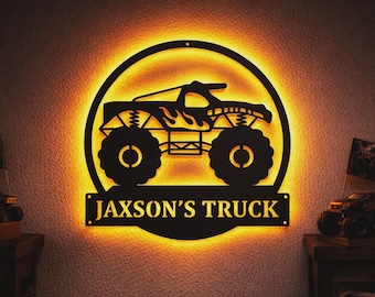 May include: Black metal monster truck sign with the text "JAXSON'S TRUCK" illuminated by a warm, yellow glow. The sign features a detailed silhouette of a monster truck within a circular frame.