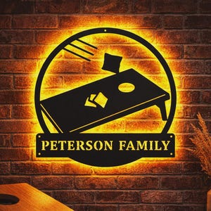 Könnte beinhalten: Schwarzes Metallschild für das Cornhole-Spiel mit dem Text "PETERSON FAMILY" an einer Backsteinmauer. Das Schild zeigt ein Cornhole-Board-Design mit einem Kreis und einem Beanbag. Das Schild wird von einem warmen, gelben Licht beleuchtet.