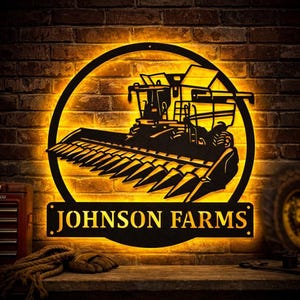 May include: A black metal sign with the words "JOHNSON FARMS" displayed on a rectangular plaque. The sign features a combine harvester silhouette within a circular frame, illuminated with a warm yellow glow against a brick wall.