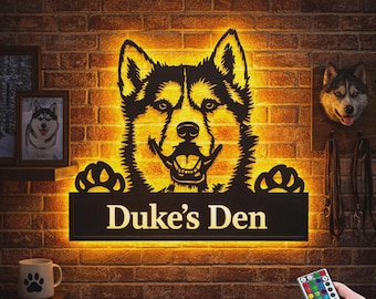 Könnte beinhalten: Ein Wand-Schild aus schwarzem Metall mit dem Scherenschnitt eines Husky-Kopfes und zwei Pfoten, die auf einer rechteckigen Platte mit der Aufschrift Duke's Den ruhen. Das Schild wird von hinten mit warmem gelbem LED-Licht beleuchtet.