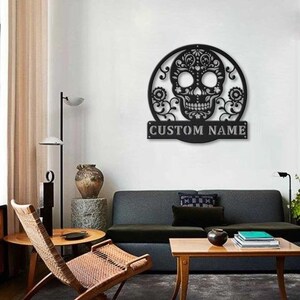 May include: Black metal wall art featuring a detailed skull design within a circular frame, with floral accents. The text "CUSTOM NAME" is displayed on a rectangular plaque below the skull. This decorative piece is suitable for interior decor.