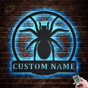 May include: A black metal spider silhouette with a circular frame and a customizable nameplate that reads "CUSTOM NAME". The spider and frame are illuminated with a bright blue light.