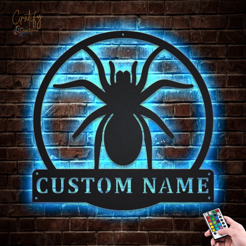 May include: A black metal spider silhouette with a circular frame and a customizable nameplate that reads "CUSTOM NAME". The spider and frame are illuminated with a bright blue light.