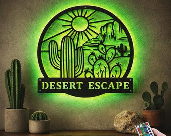May include: A circular black metal wall sign featuring a desert landscape with cacti and a sun, illuminated by a bright green LED backlight. The text "DESERT ESCAPE" is cut out in a rectangular banner at the bottom. Includes a small remote control.