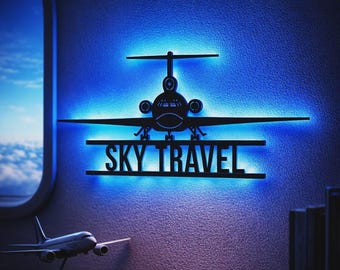 May include: A black metal wall art piece shaped like an airplane silhouette with the text SKY TRAVEL cut out below it. The design is backlit with a vibrant blue LED light, creating a glowing effect against the wall surface behind the object.