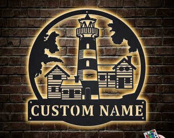 Könnte beinhalten: Schwarze Metall-Wandkunst mit einem Leuchtturm in einem runden Design mit einem Leuchtturm, zwei Häusern und einem Baum. Die Kunst hat eine personalisierte Namensschilder, auf der "CUSTOM NAME" steht.