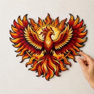 Op de afbeelding: Een wanddecoratie in de vorm van een feniks met levendige tinten rood, oranje en geel. De mythische vogel is afgebeeld met wijd gespreide vleugels, omringd door een cirkelvormig zonnestraalpatroon, alles met zwarte omlijning en gelaagd.