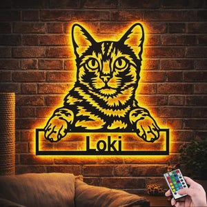 May include: A black cat-shaped wall sign with a glowing orange LED backlight. The cat is depicted from the chest up with its paws resting on a rectangular frame containing the name Loki in black lettering. The sign emits a warm orange light.
