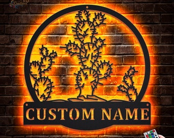 May include: A black metal silhouette of a cactus plant with a custom nameplate that reads "CUSTOM NAME". The silhouette is illuminated with warm white LED lights and is mounted on a brick wall.