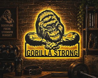 May include: A black metal wall sign featuring a gorilla with its arms crossed, illuminated by a warm yellow LED backlight. The text GORILLA STRONG is cut out at the bottom of the sign. The design is mounted on a dark brick wall surface.