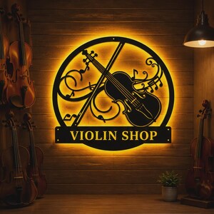 Może przedstawiać: Czarny metalowy szyld z napisem "VIOLIN SHOP" jest podświetlony na drewnianej ścianie. Znak przedstawia skrzypce, nuty i smyczek w okrągłym wzorze. Kilka skrzypiec jest widocznych w tle.