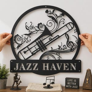 May include: A circular black metal wall sign featuring a cutout design of a trombone, musical notes, and floral swirls. Below the circle is a rectangular plaque with the text JAZZ HAVEN in bold, capital letters. The sign has two mounting holes.