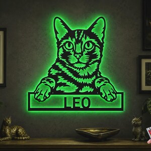 May include: A wall-mounted cat-shaped sign featuring a black silhouette of a cat with green LED backlighting. The cat is resting its paws on a rectangular frame that contains the name LEO in bold, cut-out capital letters, illuminated in green.