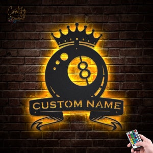 May include: Black metal wall art featuring a billiard ball with a crown on top and a banner with the text "CUSTOM NAME". The art is illuminated with warm yellow lights and comes with a remote control.