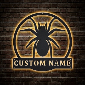May include: Black metal spider silhouette with a custom nameplate that reads "CUSTOM NAME" in a gold font. The spider is in a circle with a glowing yellow halo.