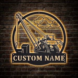 May include: Black metal sign featuring a construction crane design with the words "CUSTOM NAME" in a rectangular banner. The crane is set within a circular frame with a cloud design. The sign is illuminated with a warm, yellow glow, mounted on a brick wall.