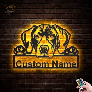 May include: A black metal silhouette of a dog's head peeking over a sign with the text "Custom Name" illuminated with warm yellow light. The sign is mounted on a brick wall. A remote control with multiple colored buttons is visible in the bottom right corner.