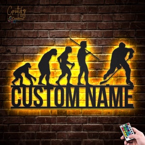 May include: Black metal wall art depicting the evolution of man, culminating in a hockey player. The art features a custom name, "CUSTOM NAME", and is illuminated with warm yellow LED lights. A remote control is included for changing the color of the lights.