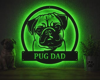 May include: A circular black metal wall sign featuring a cutout silhouette of a pug dog face. The sign is illuminated from behind with a bright green light and includes a horizontal bar at the bottom with the text PUG DAD in green letters.