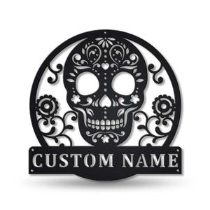May include: Black metal Day of the Dead skull wall art. The skull is intricately designed with floral patterns and set within a circular frame. The words "CUSTOM NAME" are displayed on a rectangular banner below the skull.
