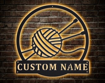 Pode incluir: Uma placa de metal preta com uma bola de lã e agulhas de tricô, dentro de uma moldura circular. A placa tem o texto "CUSTOM NAME" em uma faixa retangular. A placa é iluminada com um brilho amarelo quente, em uma parede de tijolos.