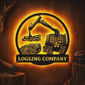 May include: A circular black metal wall sign featuring a cutout design of a logging machine lifting logs, with a stack of logs on the ground. The sign is backlit with a warm yellow glow and displays the text LOGGING COMPANY at the bottom.