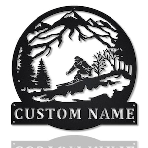 May include: Black metal wall art depicting a skier on a mountain slope, with trees and mountains in the background. The circular design includes the text "CUSTOM NAME". A decorative item for the home.