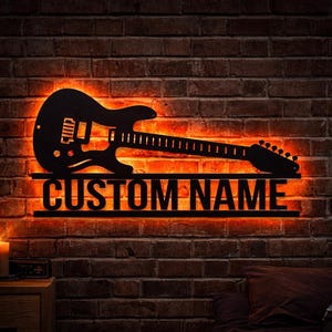 Puede incluir: Un letrero de pared negro con forma de guitarra eléctrica con el texto "CUSTOM NAME" iluminado por luces LED naranjas. El letrero está montado en una pared de ladrillo, creando un ambiente cálido y acogedor. Un gran regalo para músicos.