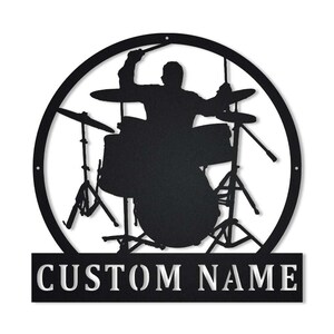 Puede incluir: Arte de pared de metal negro que representa a un baterista tocando una batería dentro de un marco circular. Las palabras "CUSTOM NAME" se muestran en una pancarta rectangular debajo. El baterista está silueteado, con las baquetas levantadas.