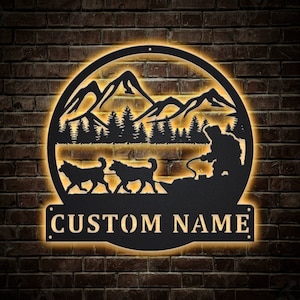 May include: Black metal silhouette of a dog sledding scene with mountains and trees in the background. The text "CUSTOM NAME" is displayed below the scene.