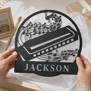 May include: A circular black metal wall sign featuring a detailed cutout of a harmonica surrounded by musical notes and staves. The name JACKSON is prominently displayed in a cutout font at the bottom of the circular frame design.