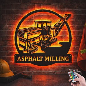 May include: A circular black metal wall sign featuring a silhouette of an asphalt milling machine with bright orange LED backlighting. The text "ASPHALT MILLING" is cut out at the bottom of the circular frame. The sign is displayed against a brick wall.