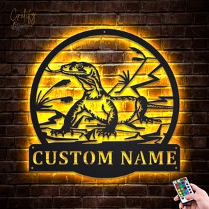 May include: A black metal wall art piece featuring a silhouette of a Komodo dragon in a circular frame with a custom nameplate. The art piece is illuminated with warm yellow LED lights and comes with a remote control for color customization.