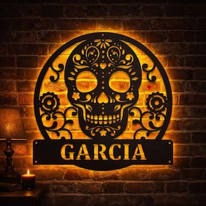 May include: A black metal Day of the Dead skull wall art with the name "GARCIA" cut out below. The skull is surrounded by a circular frame with floral designs and is illuminated by a warm, orange glow against a brick wall.