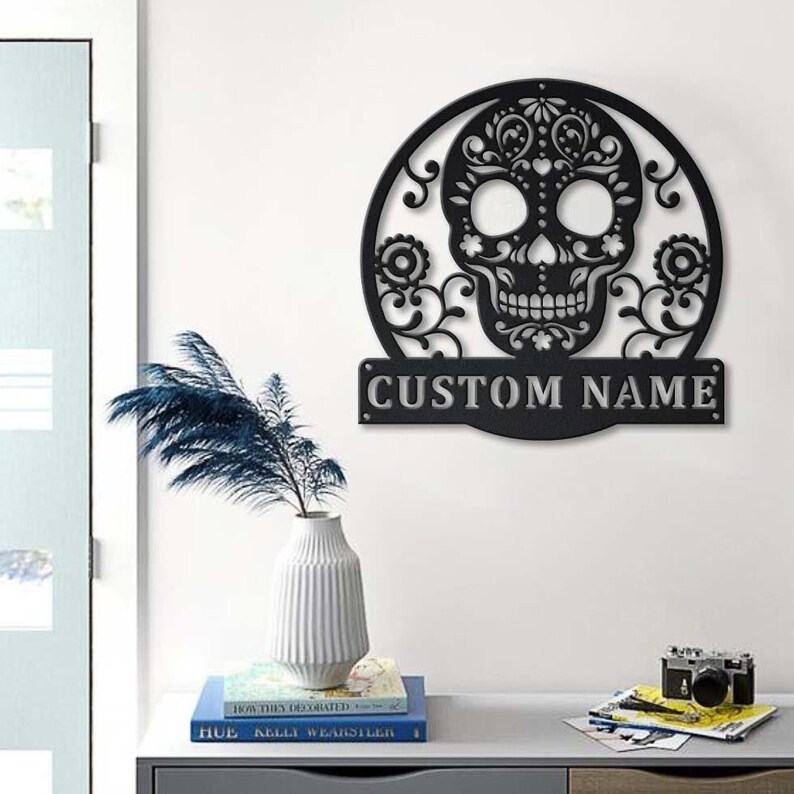 May include: Black metal Day of the Dead skull wall art with intricate floral and swirl designs. The skull is set within a circular frame and includes the text "CUSTOM NAME" on a rectangular plaque below. Decorative home decor.
