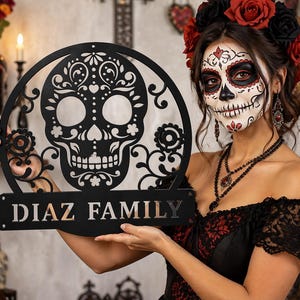 May include: A black metal Dia de los Muertos-themed family sign featuring a decorative skull design. The sign has the family name "DIAZ FAMILY" in bold letters. The skull is surrounded by floral and ornamental details.