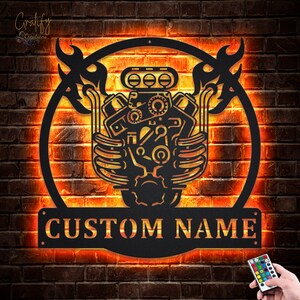 Può includere: Un decoro da parete in metallo nero con un motore stilizzato con fiamme su entrambi i lati. Il decoro ha una cornice circolare e una targhetta rettangolare con la scritta "CUSTOM NAME".