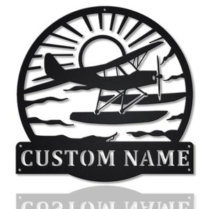 May include: Black metal silhouette of a seaplane flying over water with a sun and clouds in the background. The text "CUSTOM NAME" is on a separate black metal plaque below the silhouette.