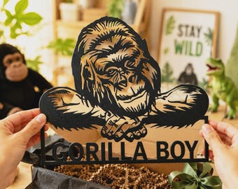 May include: A black laser-cut sign featuring a detailed illustration of a gorilla with its arms crossed. Below the gorilla, the text GORILLA BOY is cut out in bold, capital letters. The sign is held up against a blurred background.