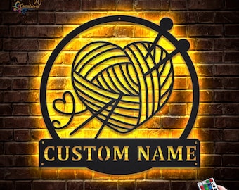 Pode incluir: Uma placa de metal preta com um novelo em forma de coração e agulhas de tricô dentro de um círculo, iluminada por uma luz amarela quente. A placa inclui o texto "CUSTOM NAME" em uma placa retangular. O fundo é uma parede de tijolos.