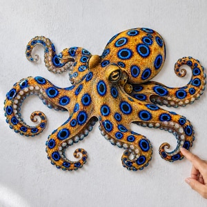 May include: A wall-mounted blue-ringed octopus sculpture featuring a textured golden-yellow body covered in vibrant, dark blue circular patterns. The octopus has eight curved tentacles extending outward, each displaying a series of suckers.